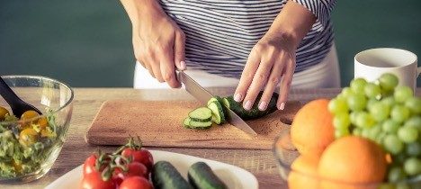 hands holding a knife cutting a cucumber on a cutting board with assorted fruits and veggies 